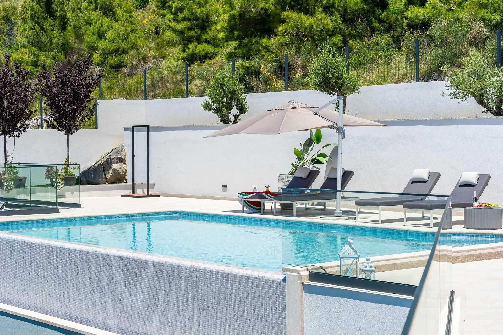 Une piscine entourée de verdure et d’un mur blanc, avec des chaises longues et des parasols.