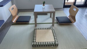 A room with a tatami mat floor, a small table with a vase, two wooden chairs with cushions, and a window with a view of the outside.