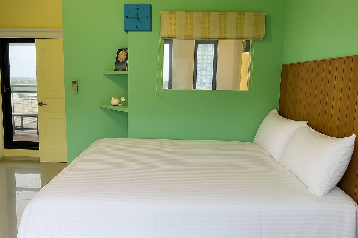 A hotel room with a large bed, a small bedside table, a clock, and a window with blinds.