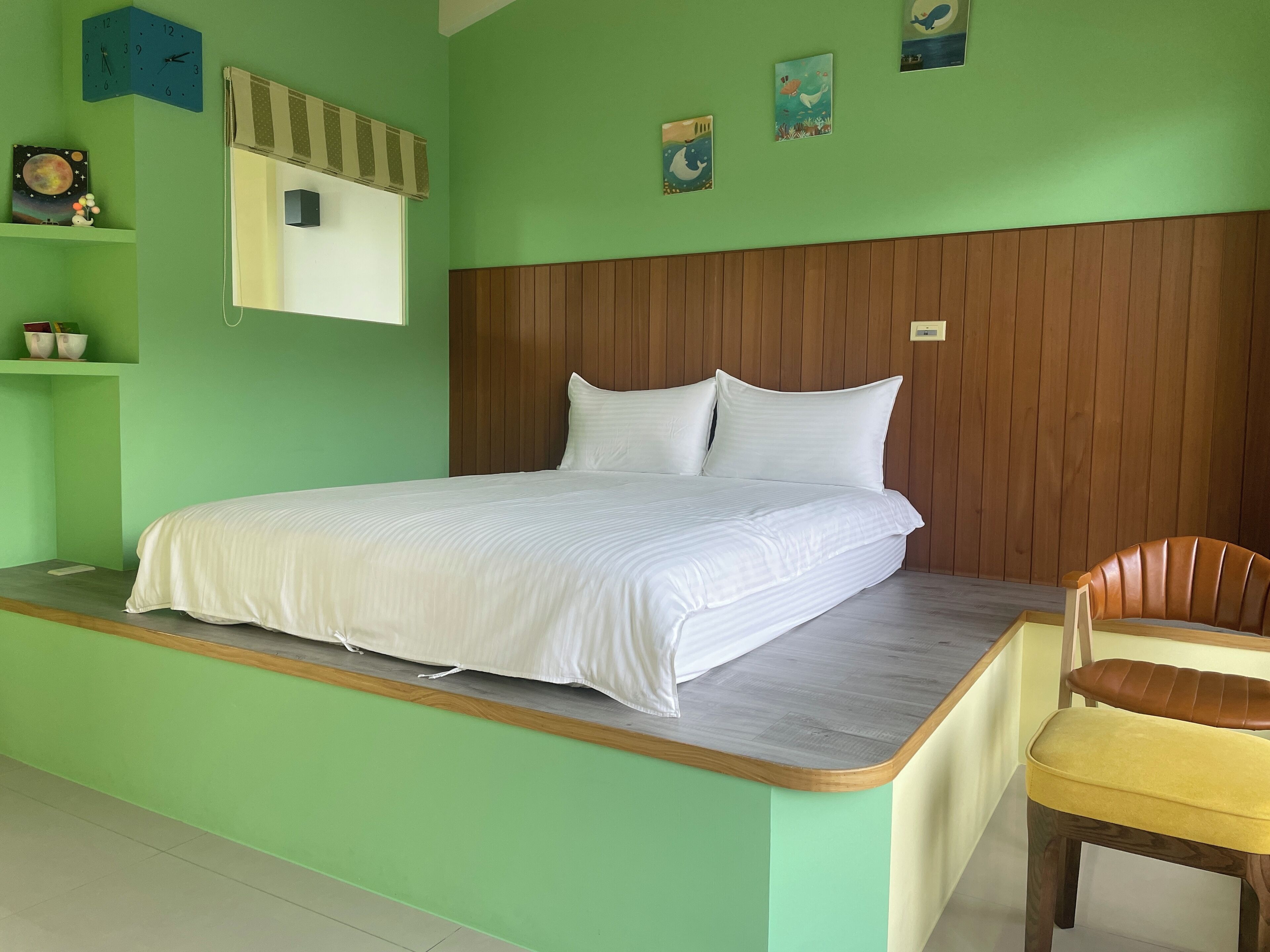 Una cama con ropa blanca, un cabecero de madera y una pared verde. Hay una silla con un cojín amarillo y un reloj en la pared.