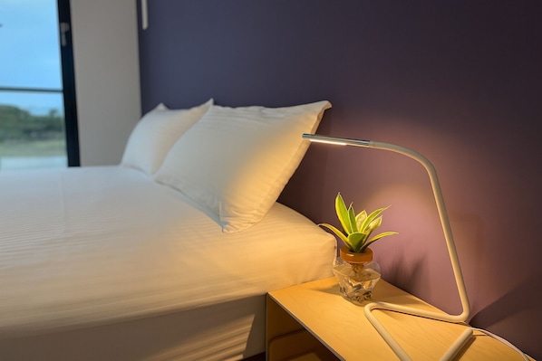 A neatly made bed with white linens, a bedside table with a lamp and a small plant, and a purple wall in the background. - Blue Sea and Sunny B&B (Toucheng)