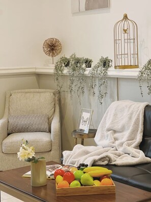 A living room with a beige armchair, a coffee table with a fruit bowl, a small side table with a picture frame, and a fireplace with hanging greenery.