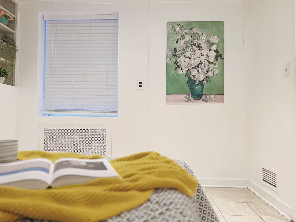 A bedroom with a bed, a window with blinds, a radiator, a painting of flowers, and a book.