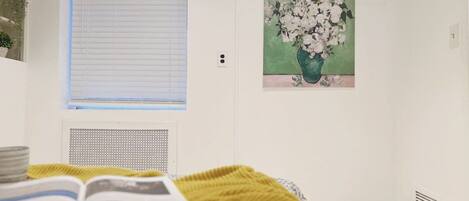 A bedroom with a bed, a window with blinds, a radiator, a painting of flowers, and a book.