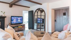 A living room with a television, a bookshelf, and a large arched window. - Luxury Villa w/ Pool, Mountain Views near NASA (Altadena)