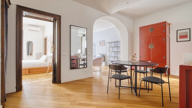 Un espacio habitable moderno con suelo de madera, una zona de comedor con mesa y sillas, un armario rojo y un dormitorio con cama y espejo.