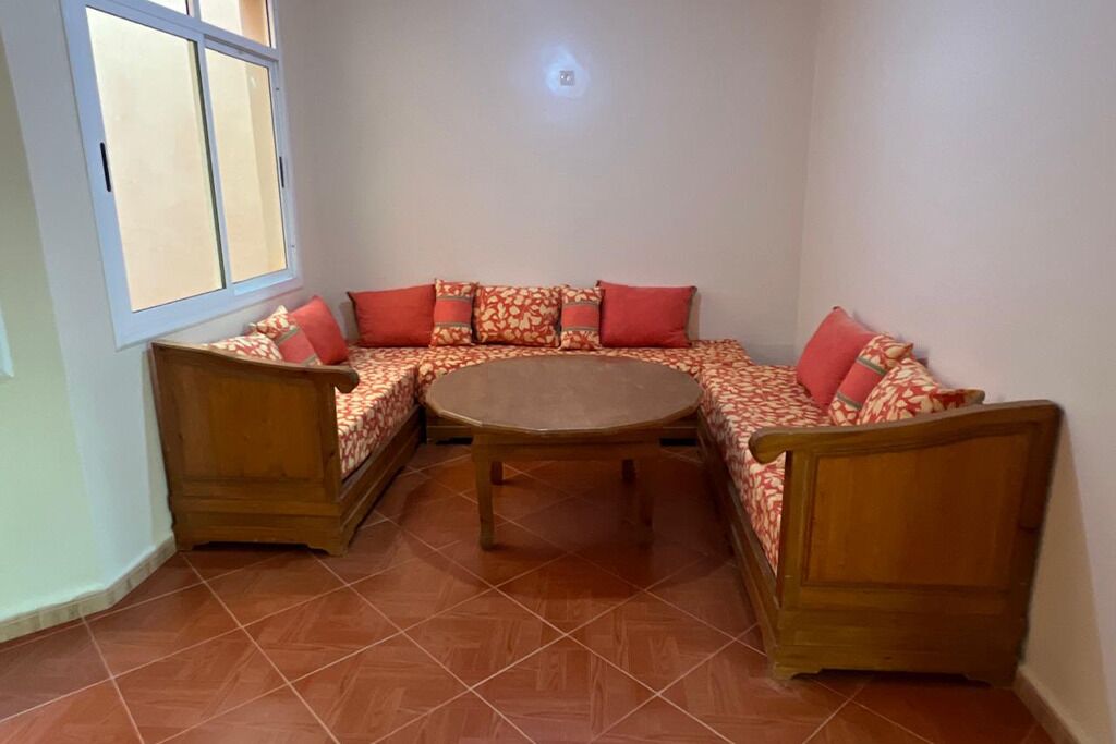 Habitación de esquina con un banco de madera y una mesa redonda, una ventana y una lámpara fijada a la pared.