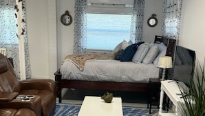 A cozy living room with a brown leather sofa, a white coffee table, and a bed with a view of the ocean.