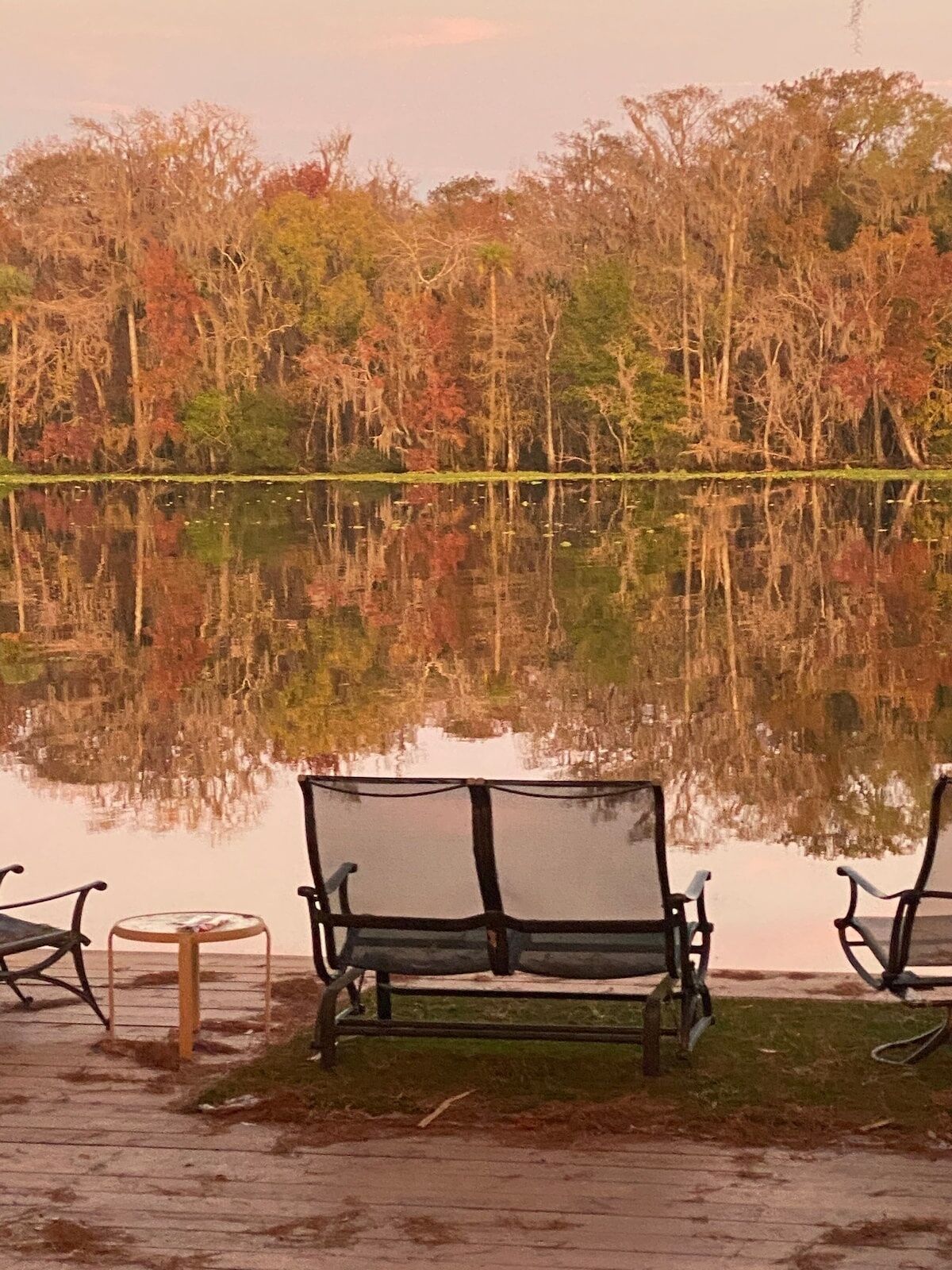 A bench and chairs by a calm lake with trees in the background.