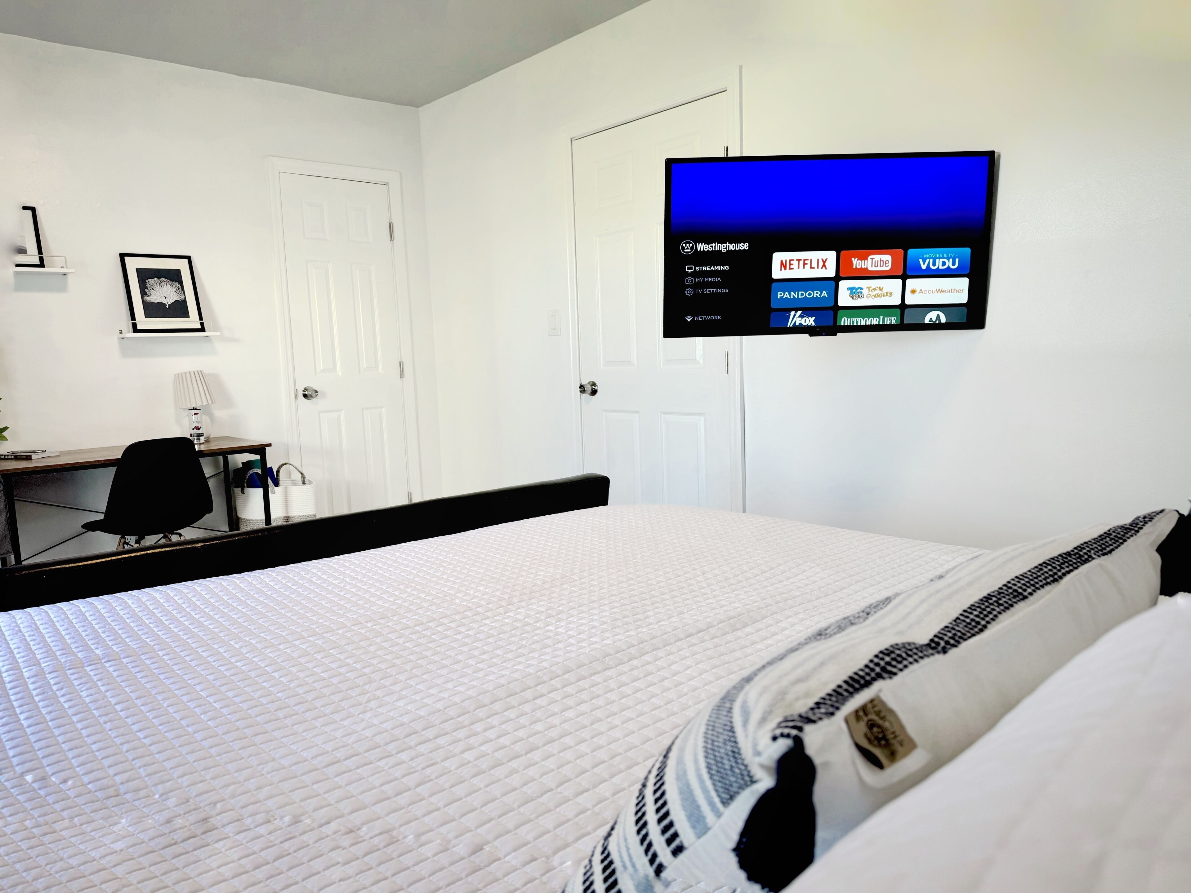 A hotel room with a bed, a desk, a chair, a TV mounted on the wall displaying various streaming services, and a door.