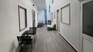 A narrow corridor with a dining area featuring a table and chairs, a sofa, and a window with blinds.