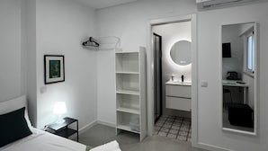 A modern hotel room with a neatly made bed, a bedside lamp, a wardrobe, a bathroom with a mirror, and an air conditioning unit.