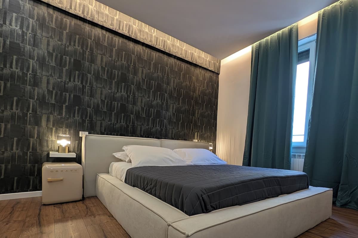Un dormitorio moderno con una cama grande, una mesita de noche, una lámpara de pared y una ventana con cortinas.