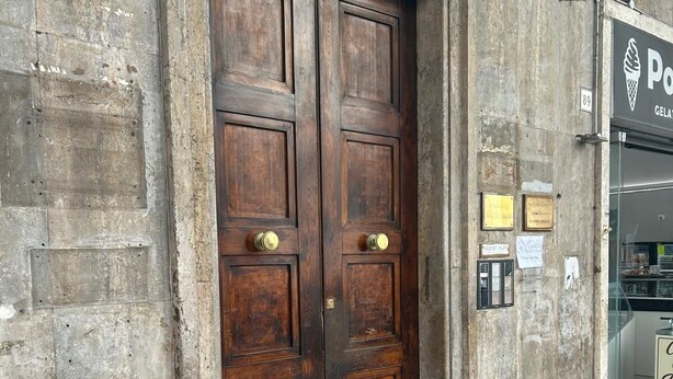 Una amplia puerta doble de madera con pomos de latón, instalada en un edificio de concreto con piso de baldosas y un diseño blanco y rojo con motivos.