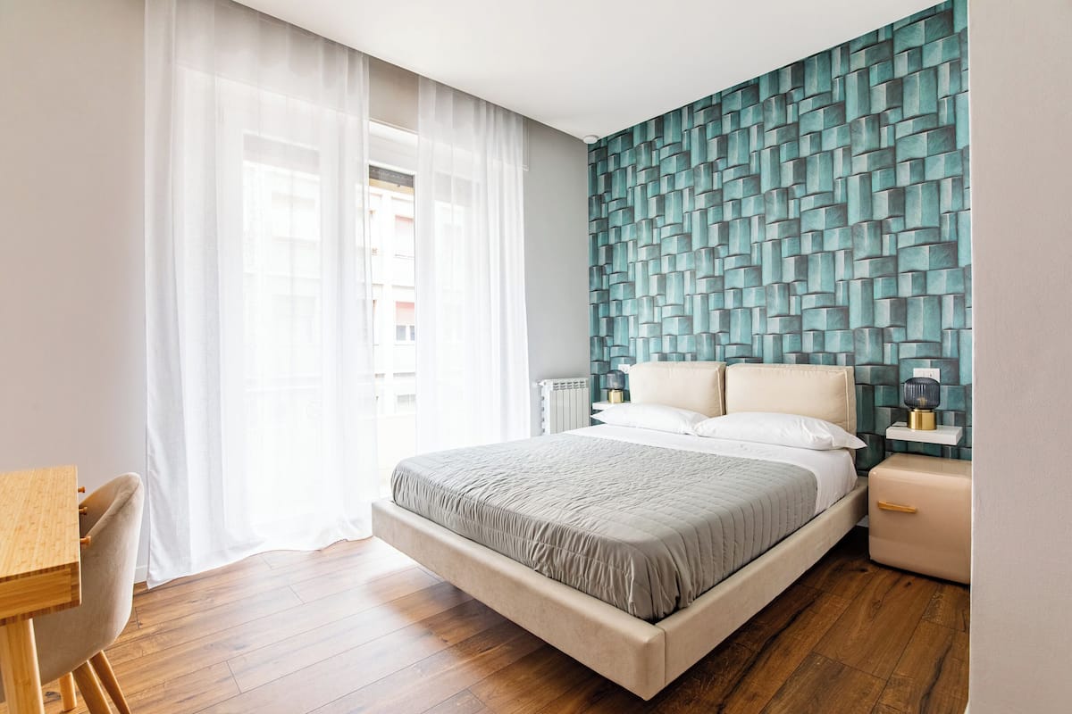 Un dormitorio moderno con una cama grande, un escritorio y vistas de la ciudad a través de cortinas translúcidas.