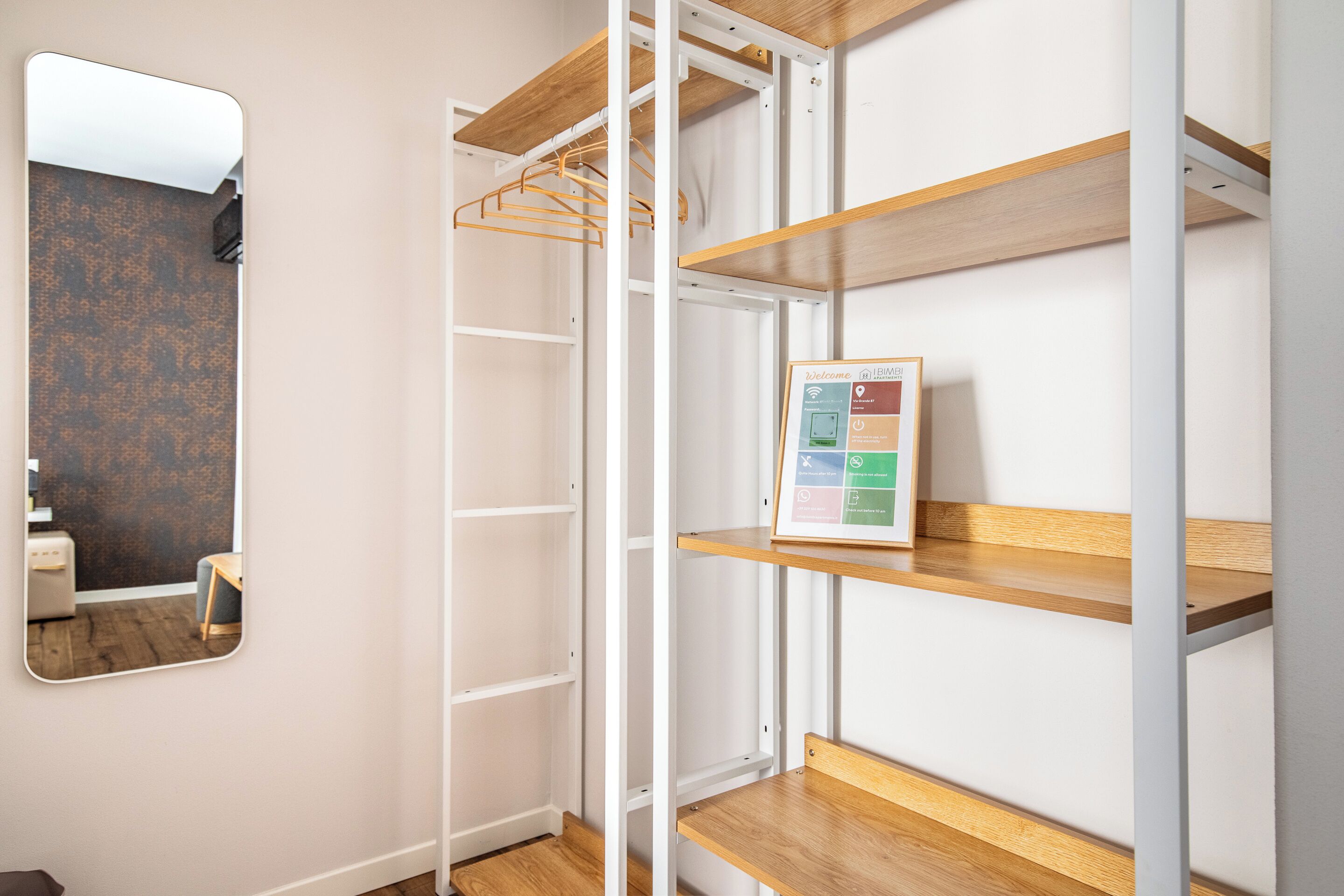 A room with a wooden floor, a white wall, and a mirror. There are shelves with a welcome sign and a small table.