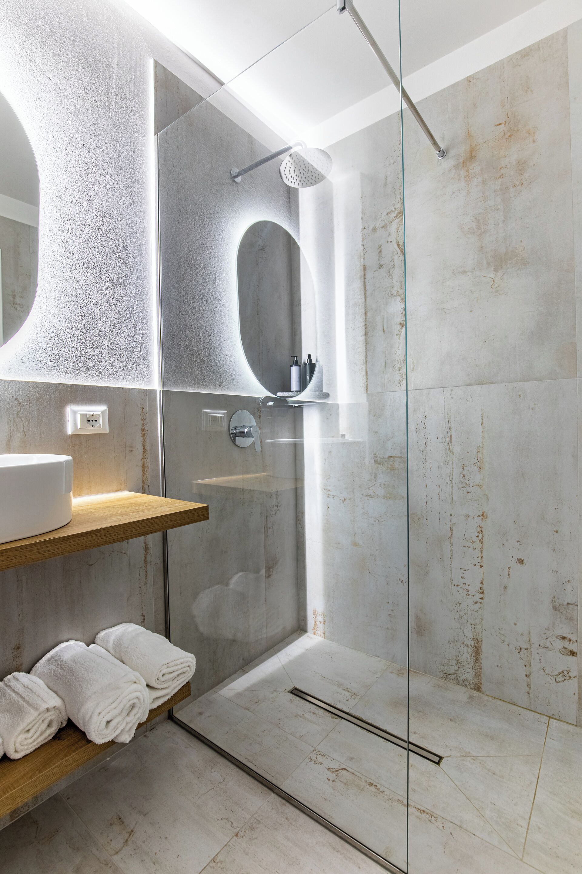 A modern bathroom with a glass-enclosed shower, a wooden shelf with towels, and a large mirror.