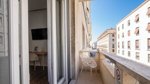 Un balcón con una silla y una mesa, una puerta que da a una habitación y vistas a edificios.