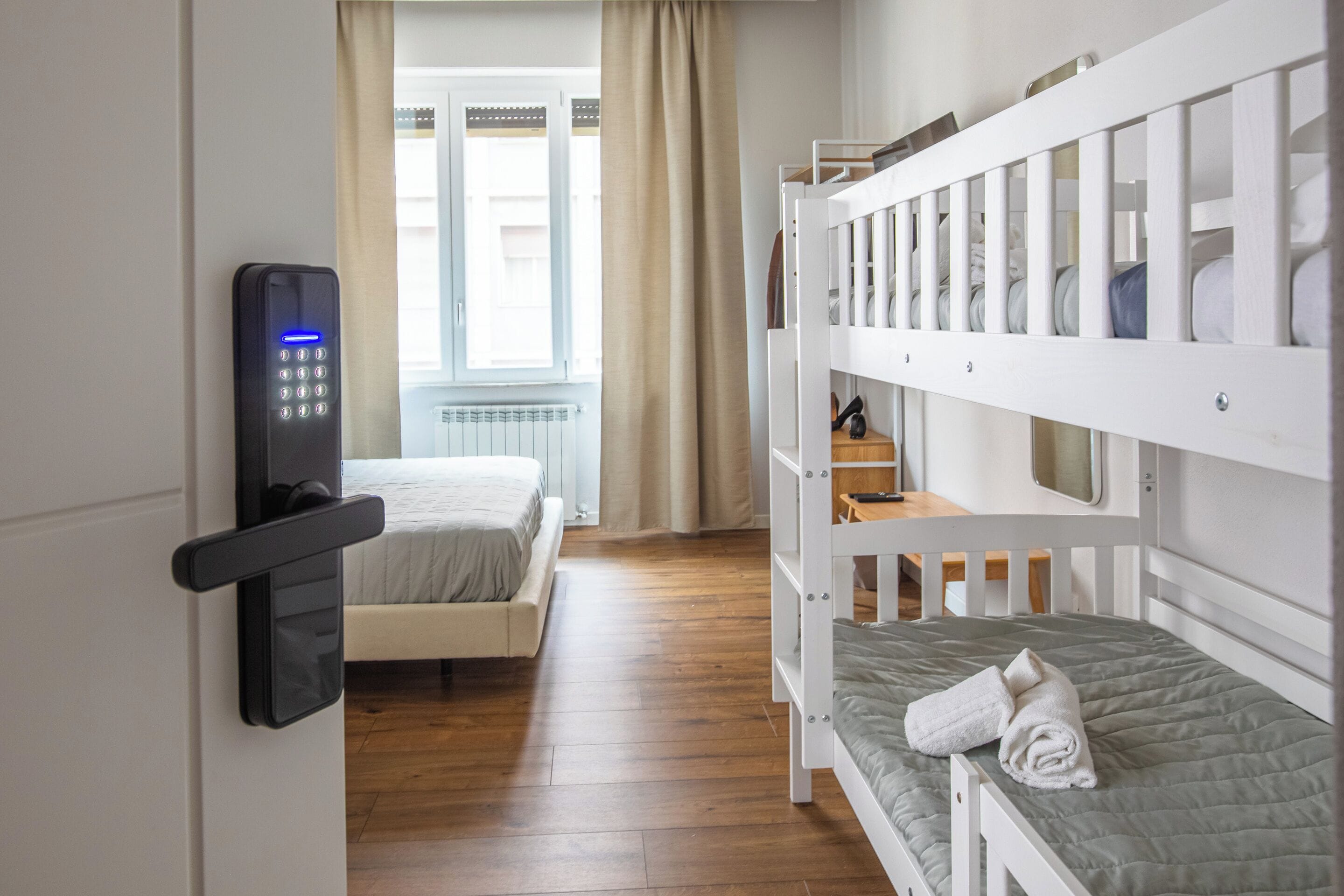 Ein Hotelzimmer mit Etagenbett, einem Einzelbett, Holzboden, einem Fenster mit Vorhängen und einem digitalen Schloss an der Tür.
