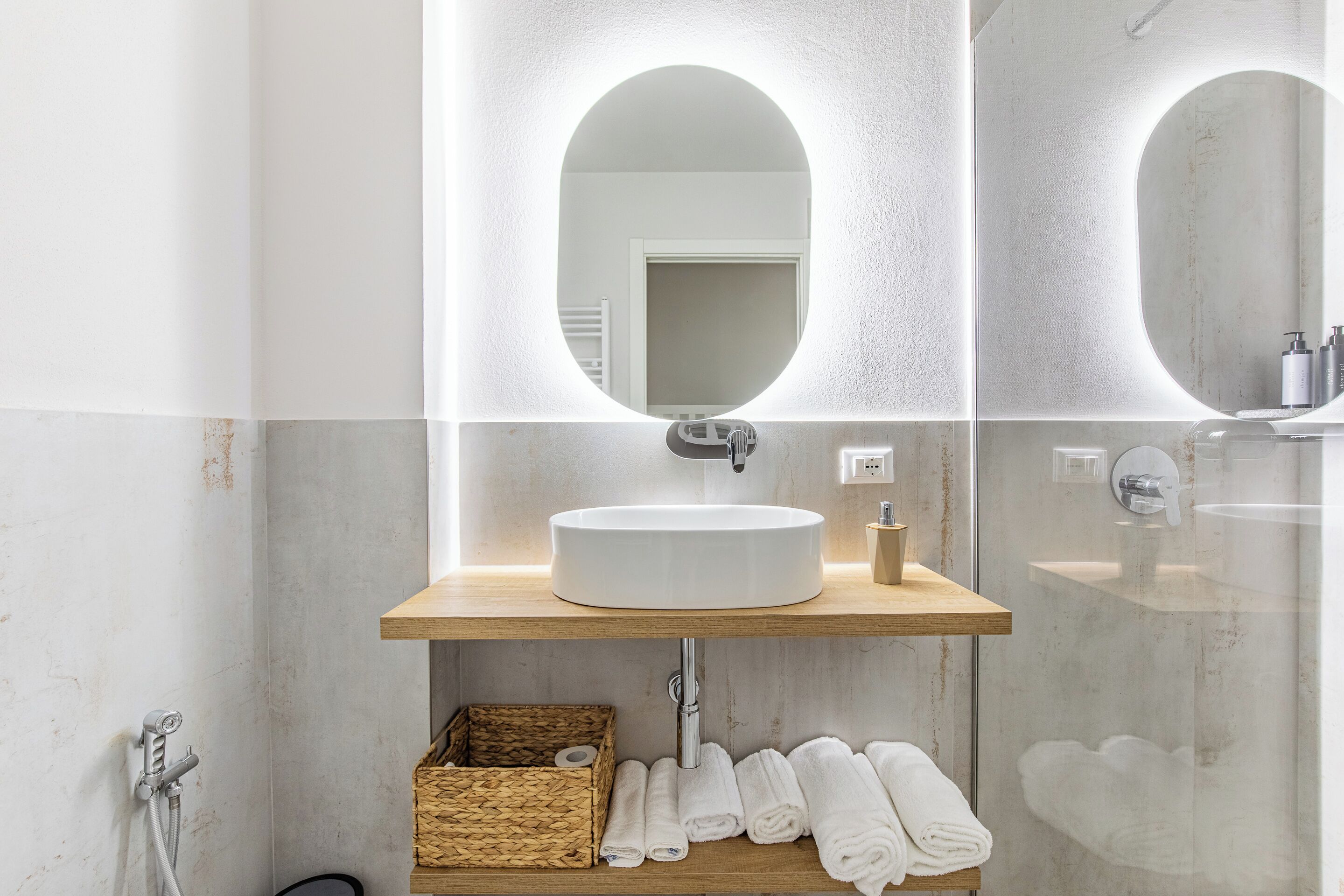 A bathroom with a round mirror, a white sink, and a wooden shelf with towels and a basket.