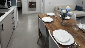 A modern kitchen with white cabinets, a wooden dining table set for four, and a living area with a sofa and a decorative wall piece.