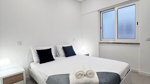 A modern bedroom with a large bed, white walls, a window, and a nightstand.