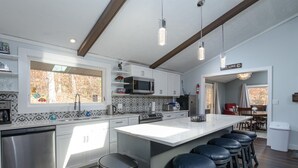 A modern kitchen with white cabinets, a large island with bar stools, stainless steel appliances, and a view of the outdoors. - Mountain side, remodeled  4 br neat trail head. (Wilmington)
