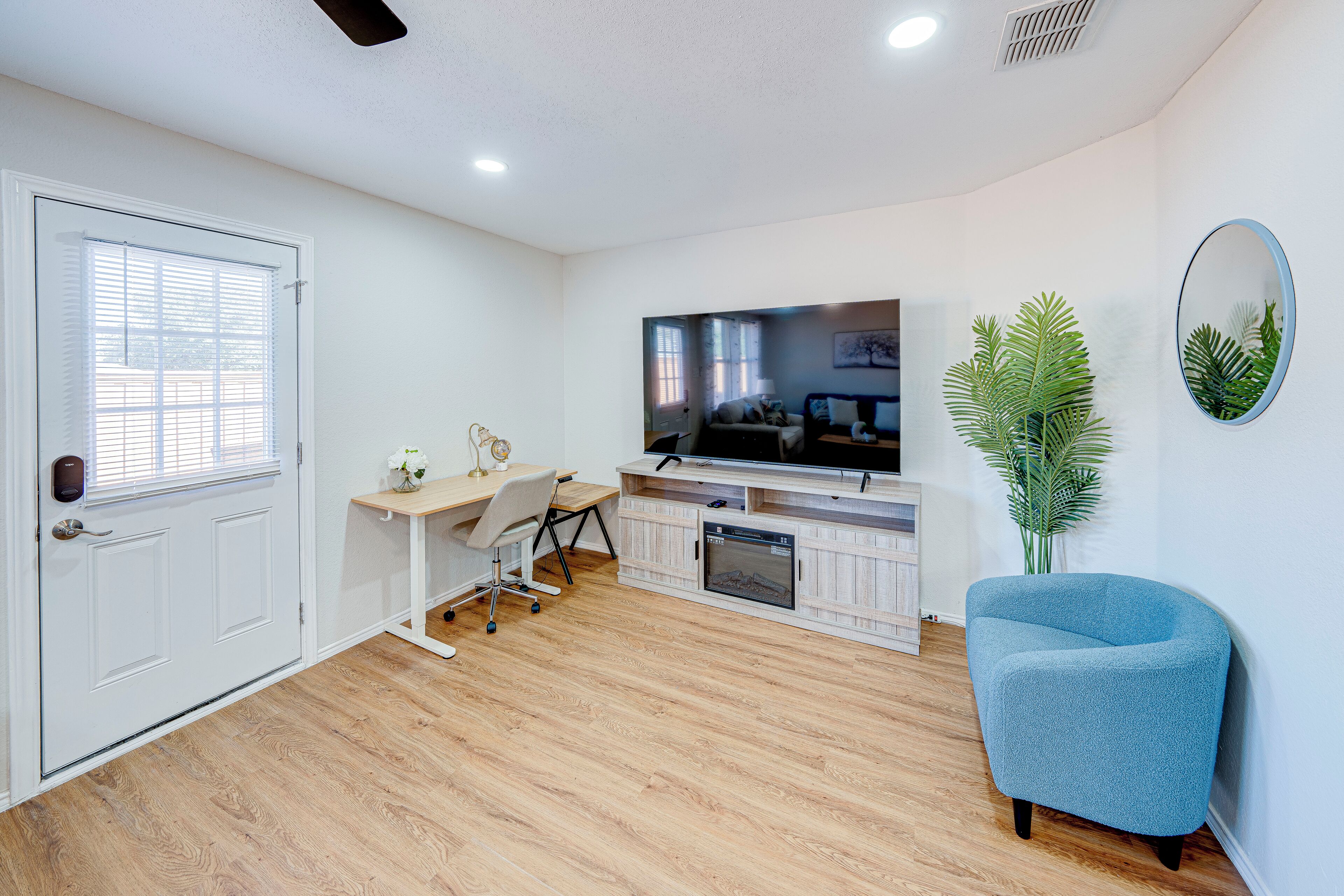 A modern living room with a flat-screen TV, a blue armchair, a wooden floor, a white door, and a small desk with a chair.