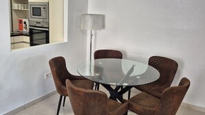 A dining area with a glass table and four brown upholstered chairs. A lamp is beside the table, and a kitchen is visible through an open door.