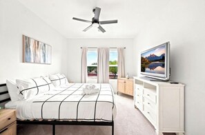 Een slaapkamer met een bed, een televisie, een plafondventilator, een raam met gordijnen en een schilderij aan de muur.