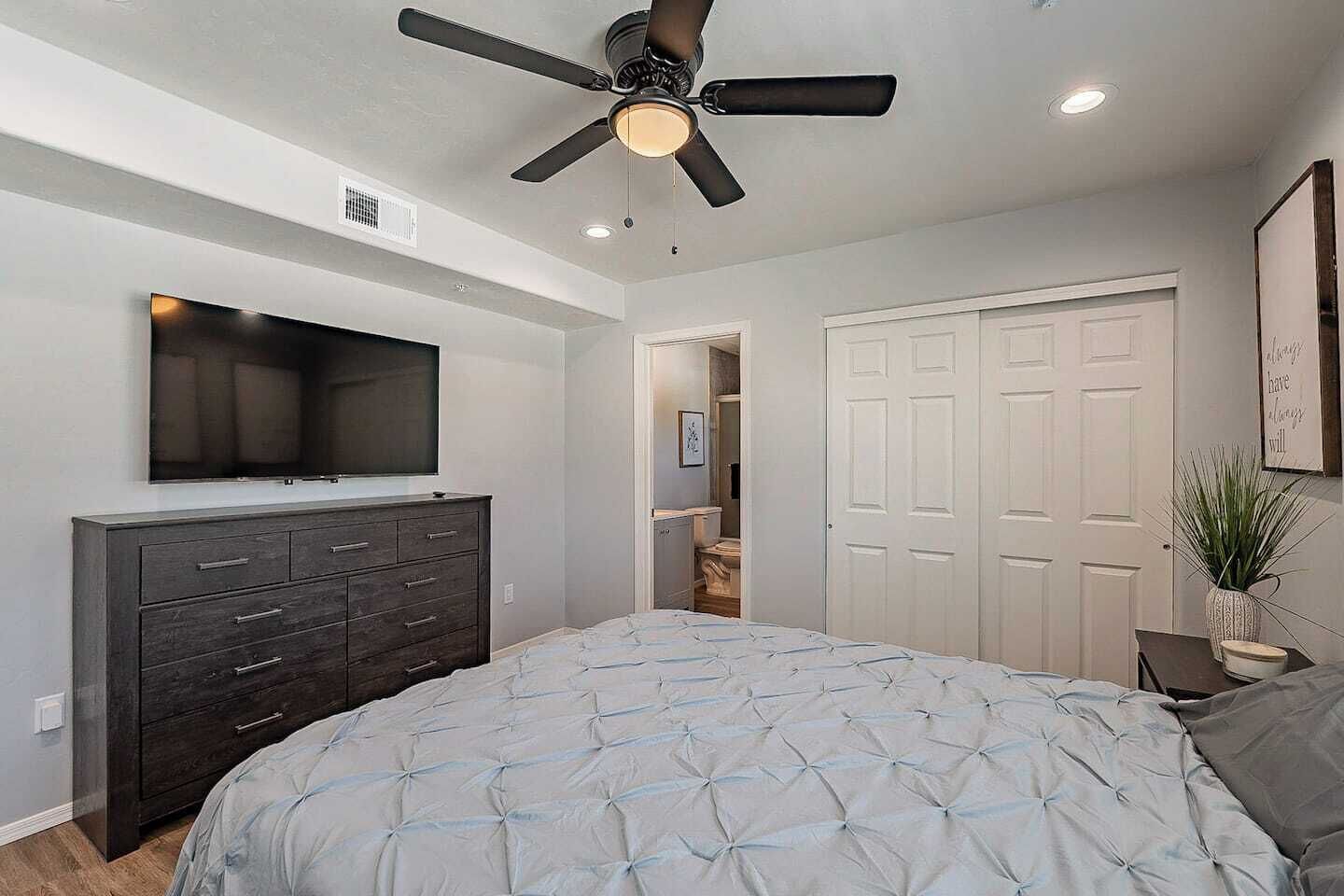 A bedroom with a bed, a dresser, a ceiling fan, a TV, and a closet.