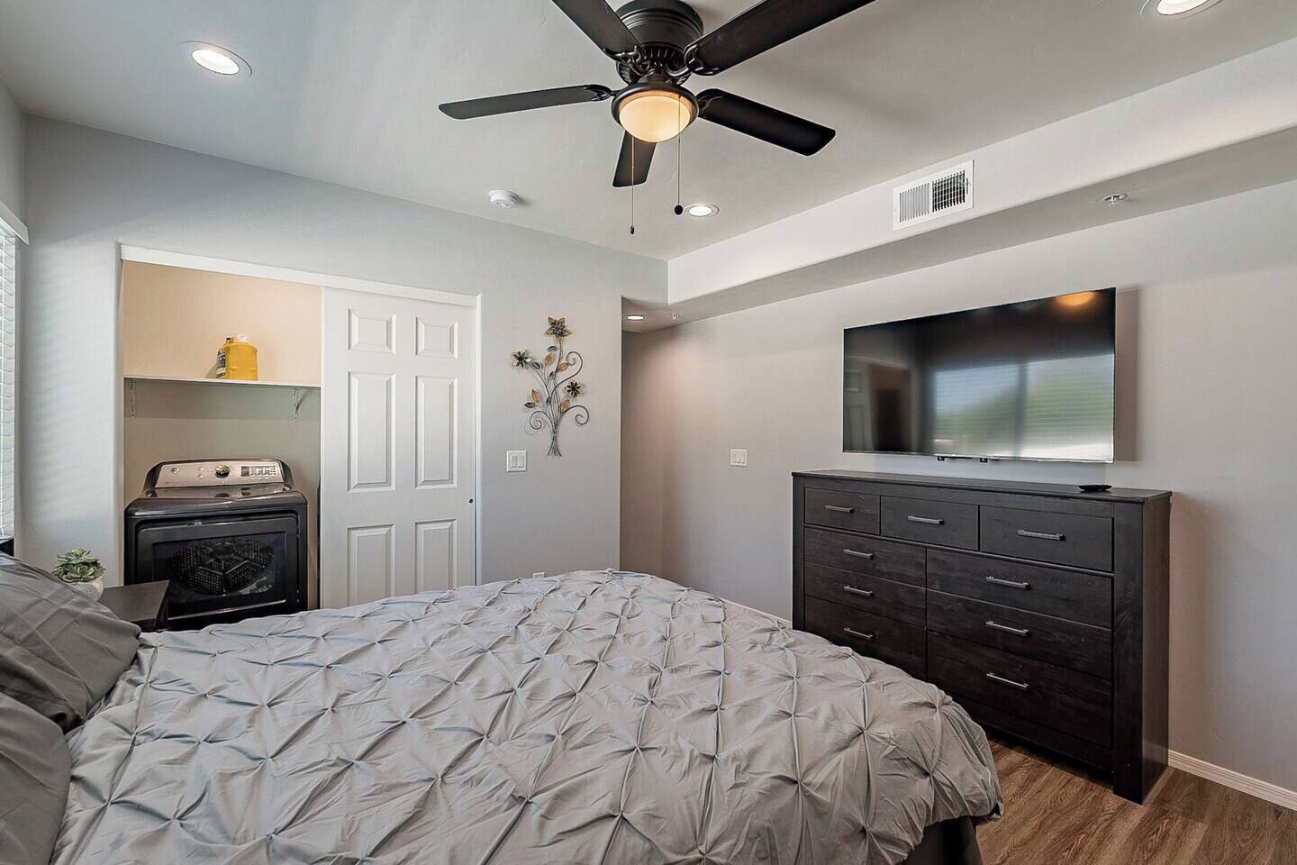 A bedroom with a bed, dresser, ceiling fan, and a TV on the wall.