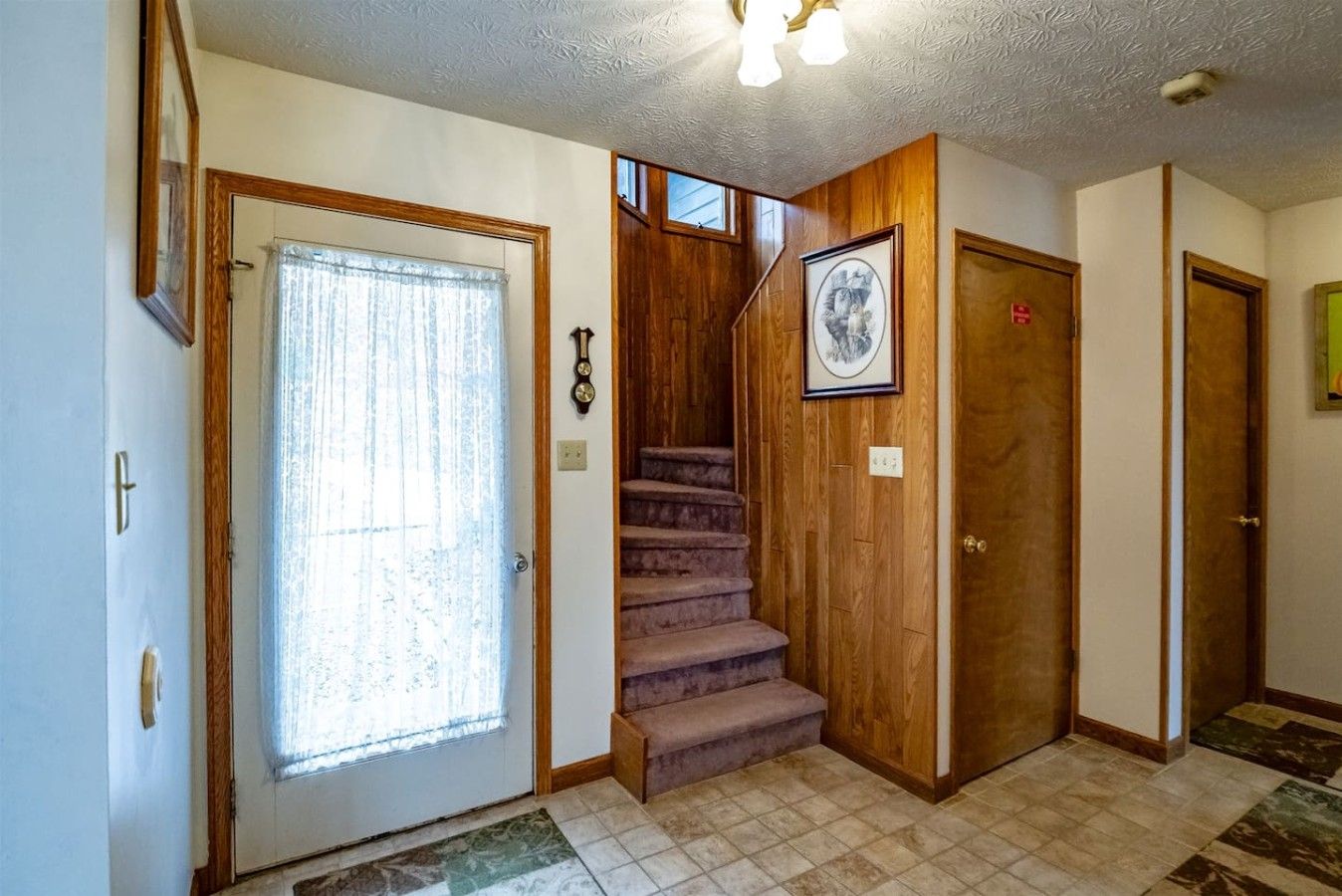 A hallway with a wooden door, a window with a curtain, a staircase, and a framed picture on the wall.