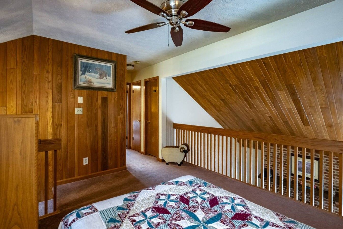 A bedroom with a wooden ceiling, a bed with a patterned quilt, a ceiling fan, and a door leading to another room.