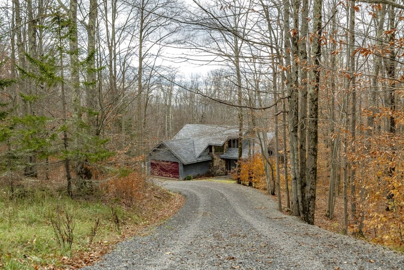 A rural road leading to a stone house with a red garage door, surrounded by trees and a gravel path.
