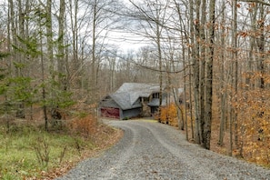 A rural road leading to a stone house with a red garage door, surrounded by trees and a gravel path.