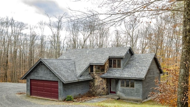A two-story wooden cabin with a red garage door, surrounded by a forested area.
