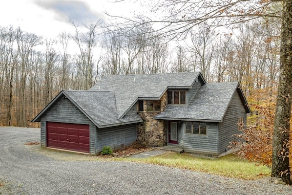 A two-story wooden cabin with a red garage door, surrounded by a forested area.