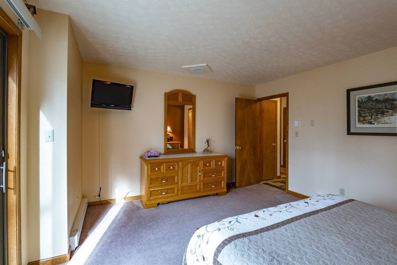 Quarto com cama, cômoda de madeira, televisão fixada na parede e porta que dá para outro espaço.