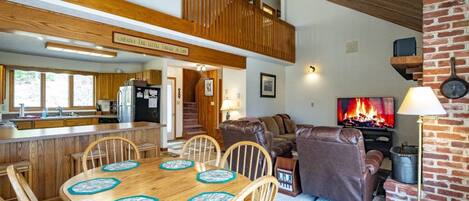 A spacious living area with a fireplace, a dining table with chairs, and a kitchen in the background.