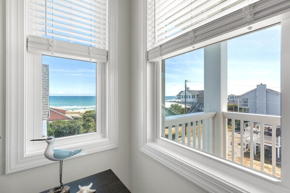 Habitación con vistas a la playa y al océano a través de ventanas grandes con persianas blancas.