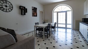 A spacious living area with a dining table, chairs, a sofa, a large wall clock, and a kitchen area with a tiled floor and a window.