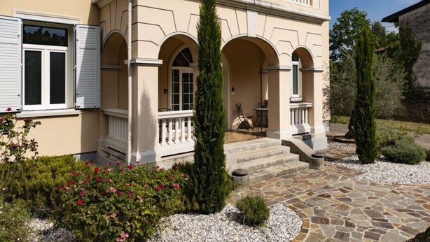 A two-story building with a balcony, stone steps, and a garden with shrubs and flowers.