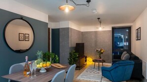 A modern living room with a dining area, featuring a round mirror, a wooden dining table with fruit, a blue chair, and a black sofa. - Kula Manchester (MANCHESTER)