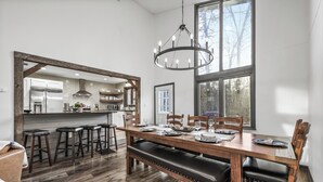 A modern dining area with a wooden table, bar stools, and a bench. A large mirror reflects the kitchen area. - 3,800 Sq Ft Luxury Cabin on Private Acre, Hot Tub, 2 Game Rooms,  Sleeps 16 (Sevierville)