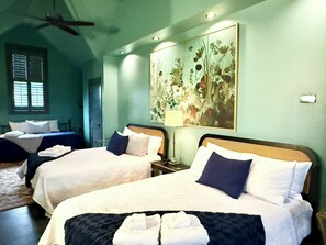 Une chambre d’hôtel avec deux lits, un ventilateur de plafond, un grand tableau représentant des motifs floraux et une fenêtre avec des stores.