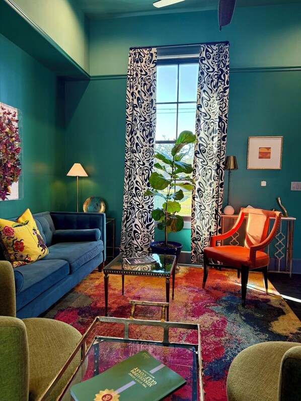 Un salon aux murs de couleur turquoise, avec un canapé bleu, un fauteuil orange, une table basse en verre et un tapis aux couleurs vives.