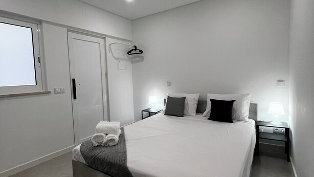 A modern bedroom with a large bed, a bedside table with a lamp, a hanging coat rack, and a window with a view.