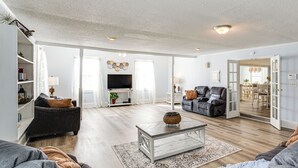 Ein geräumiges Wohnzimmer mit grauem Sofa, Couchtisch und Fernseher. Durch eine offene Tür ist ein Essbereich sichtbar.