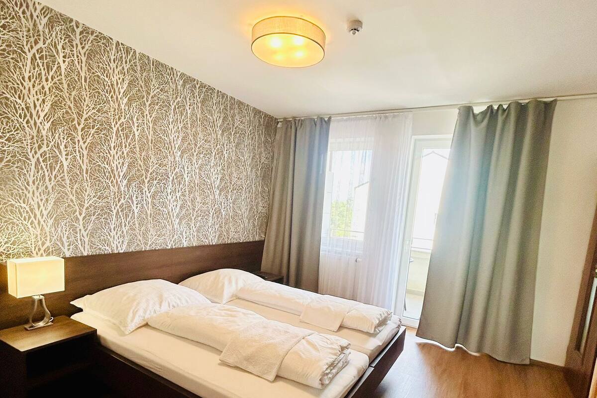 A hotel room with a wooden floor, a bed with white linens, a nightstand with a lamp, and a patterned wall.